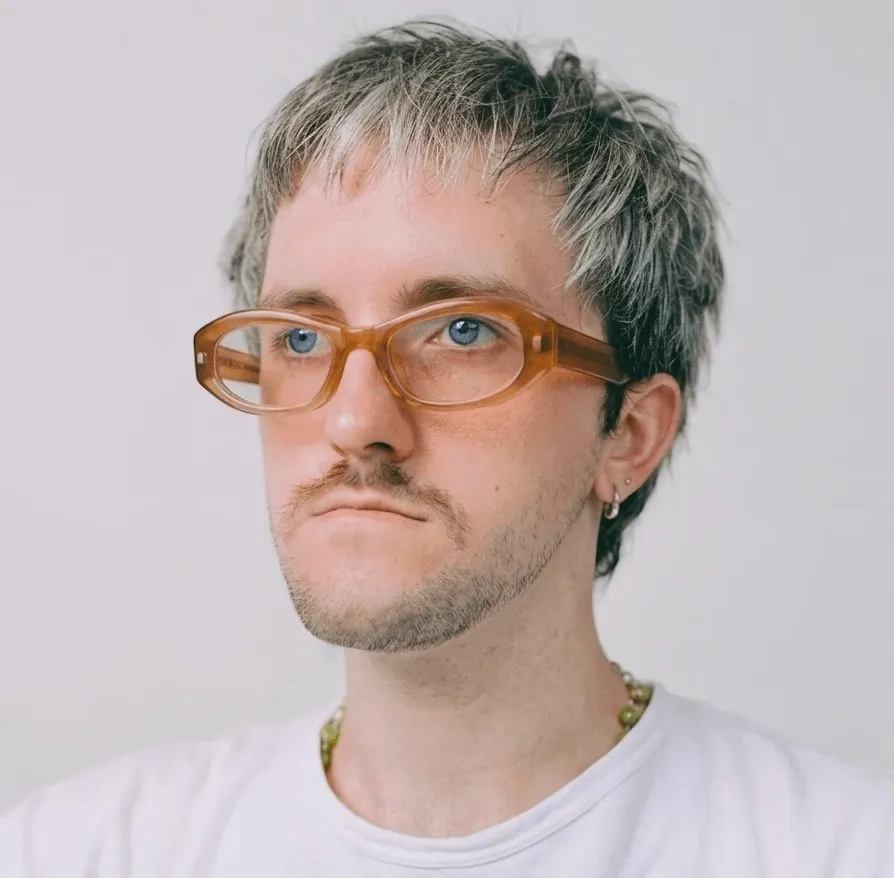 Portrait of Tom Le Masson Banning Lover: short grey hair, amber glasses and a white T-shirt, looking off to the side.